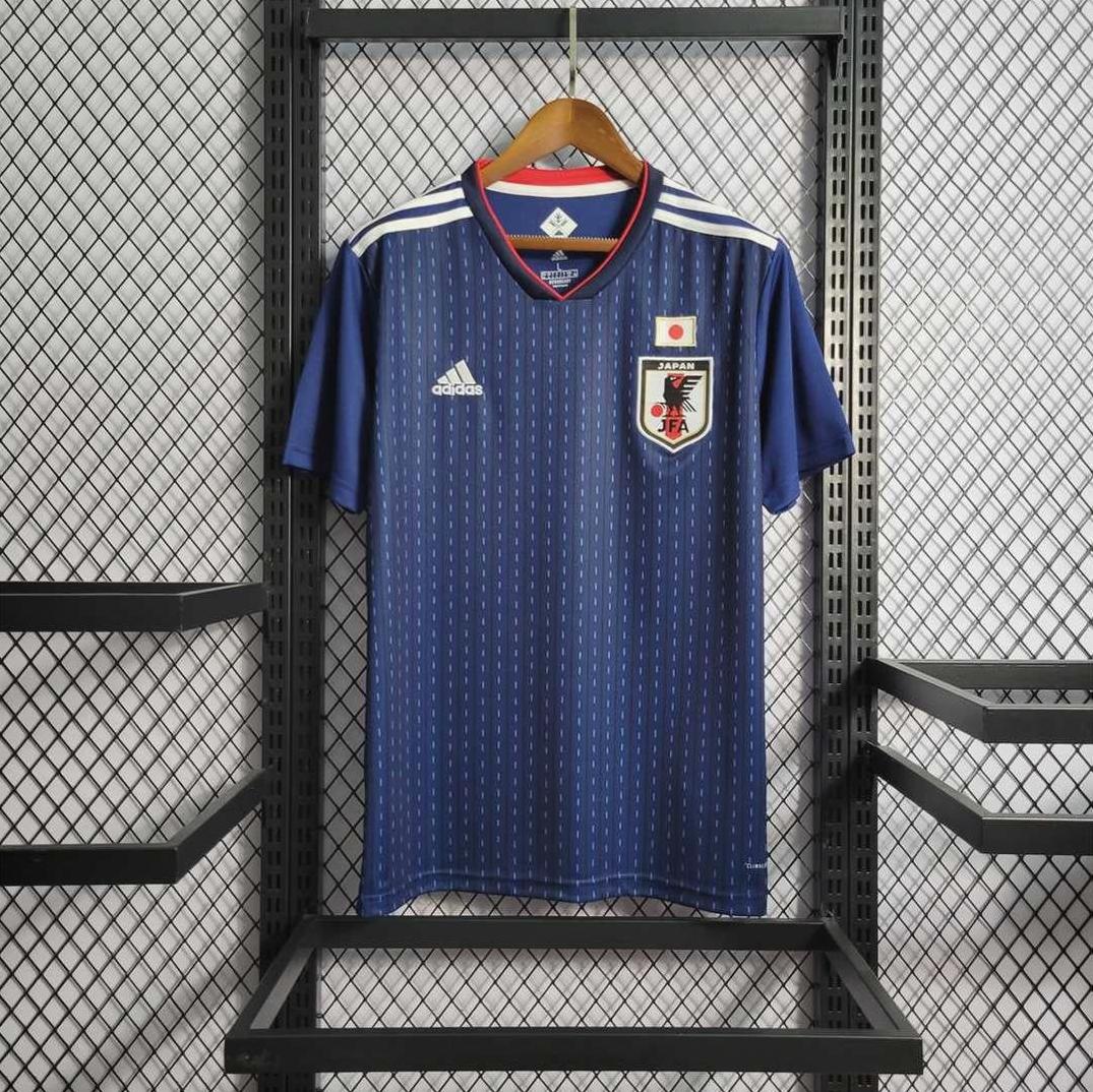 A Japan 2018 Home Shirt by Retro1999, in blue with white and red accents, hangs on a wooden hanger against a metal mesh background. The kit displays the team crest, Adidas logo, and a small Japanese flag to showcase national pride.