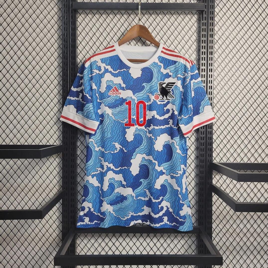 A blue, white, and red Japan 2022 Special Edition jersey by Retro1999, with wave patterns, number 10, bird logo, and Japanese flag patch hangs on a wooden hanger against a metal grid wall.
