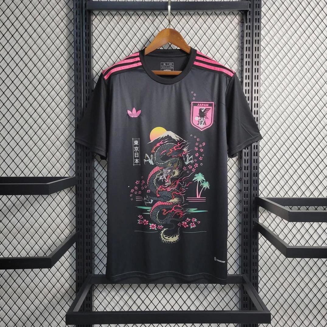 A black Japan 2023 Black Training Shirt by Retro1999 hangs on a wooden hanger, featuring pink accents, a dragon graphic, rising sun, Japanese motifs, pink Adidas logo, and Japan’s crest against a metal grid backdrop.