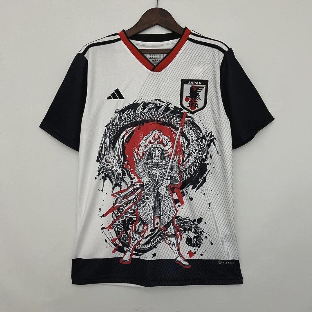 A white and black Retro1999 Japan 2023 Special Edition soccer jersey hangs on a wooden hanger, featuring an illustrated red-armored samurai with a sword before a black dragon, plus the Japan football crest on the chest.