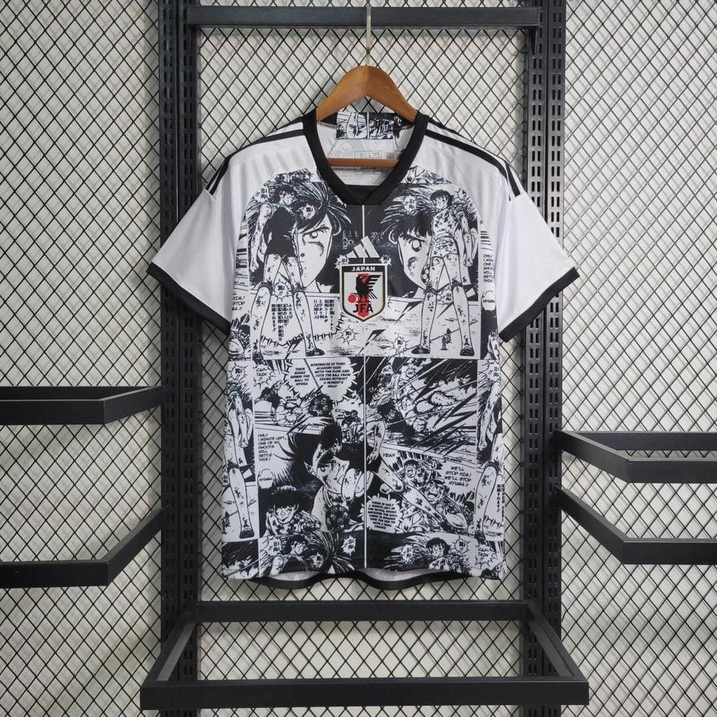 The Retro1999 Japan 2023/24 Anime Special Edition jersey features black and white manga-style graphics and a Japan football emblem, displayed on a wooden hanger against a metal mesh background.