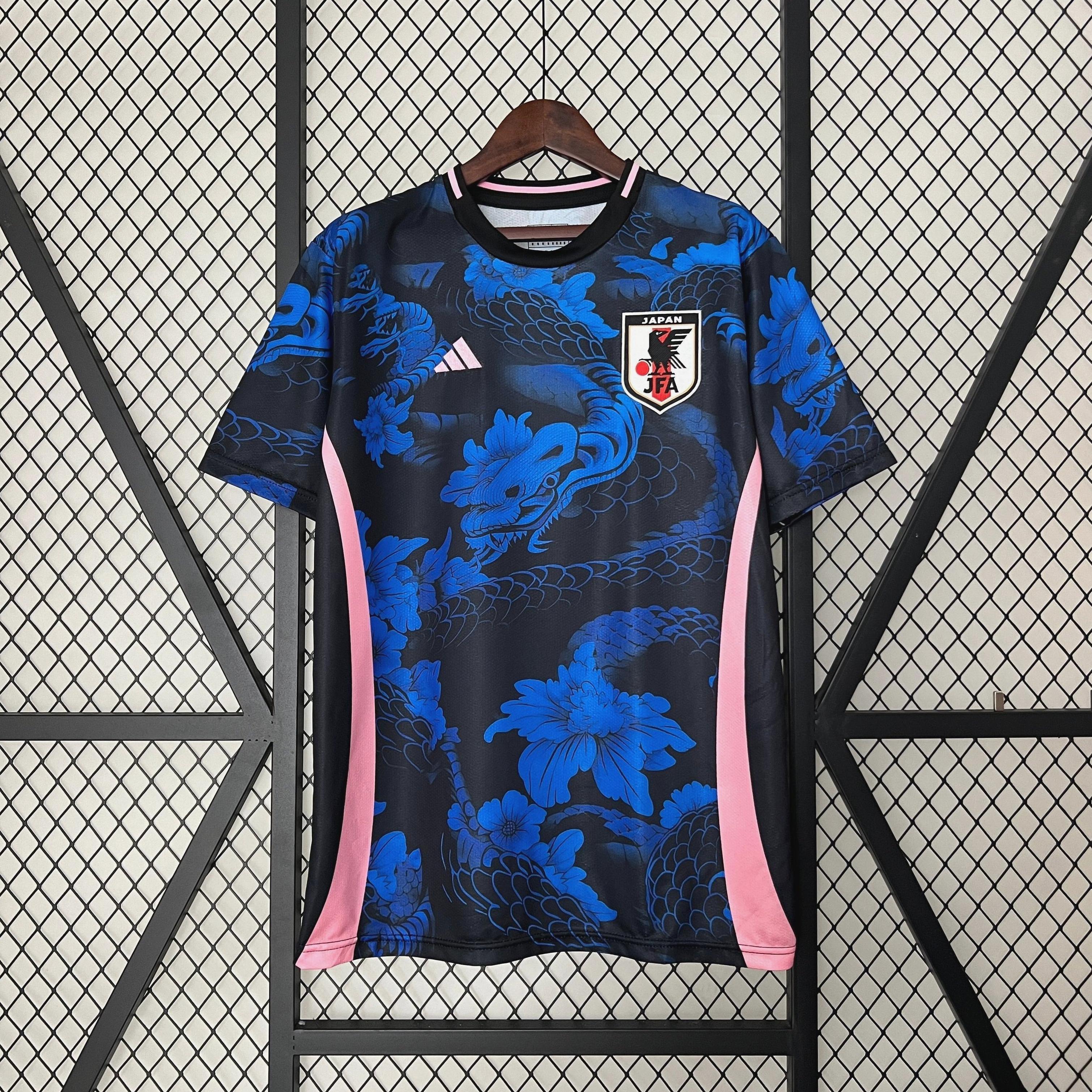 The Retro1999 Japan 2024-25 Special Edition soccer jersey, featuring blue and black dragon and floral patterns, pink side stripes, and the national crest, hangs on a wooden hanger against a metal grid.