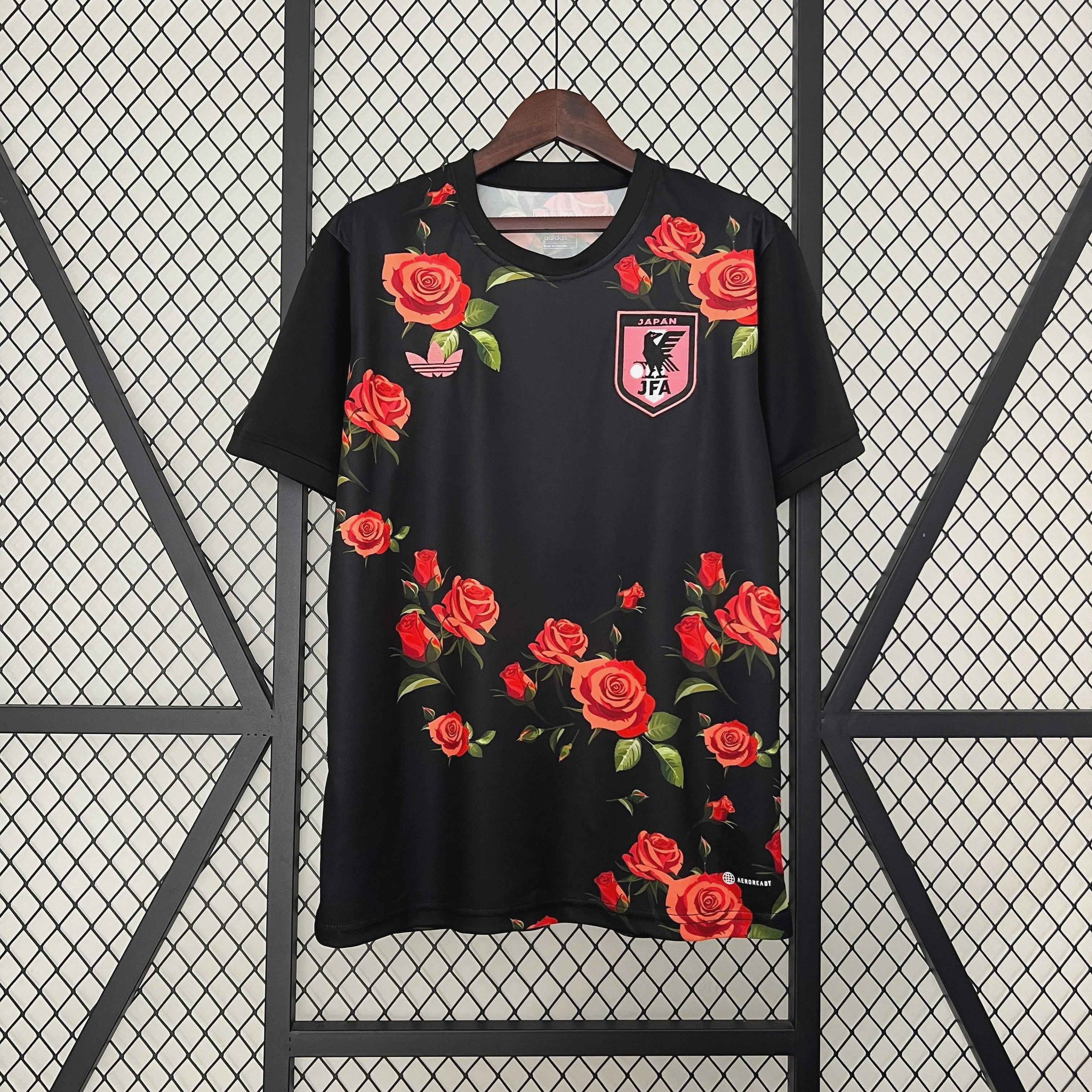 The Retro1999 Japan 2024 Roses Special Edition black soccer jersey displays red rose patterns and a pink badge on the chest, hanging on a wooden hanger against a metal wire grid background.