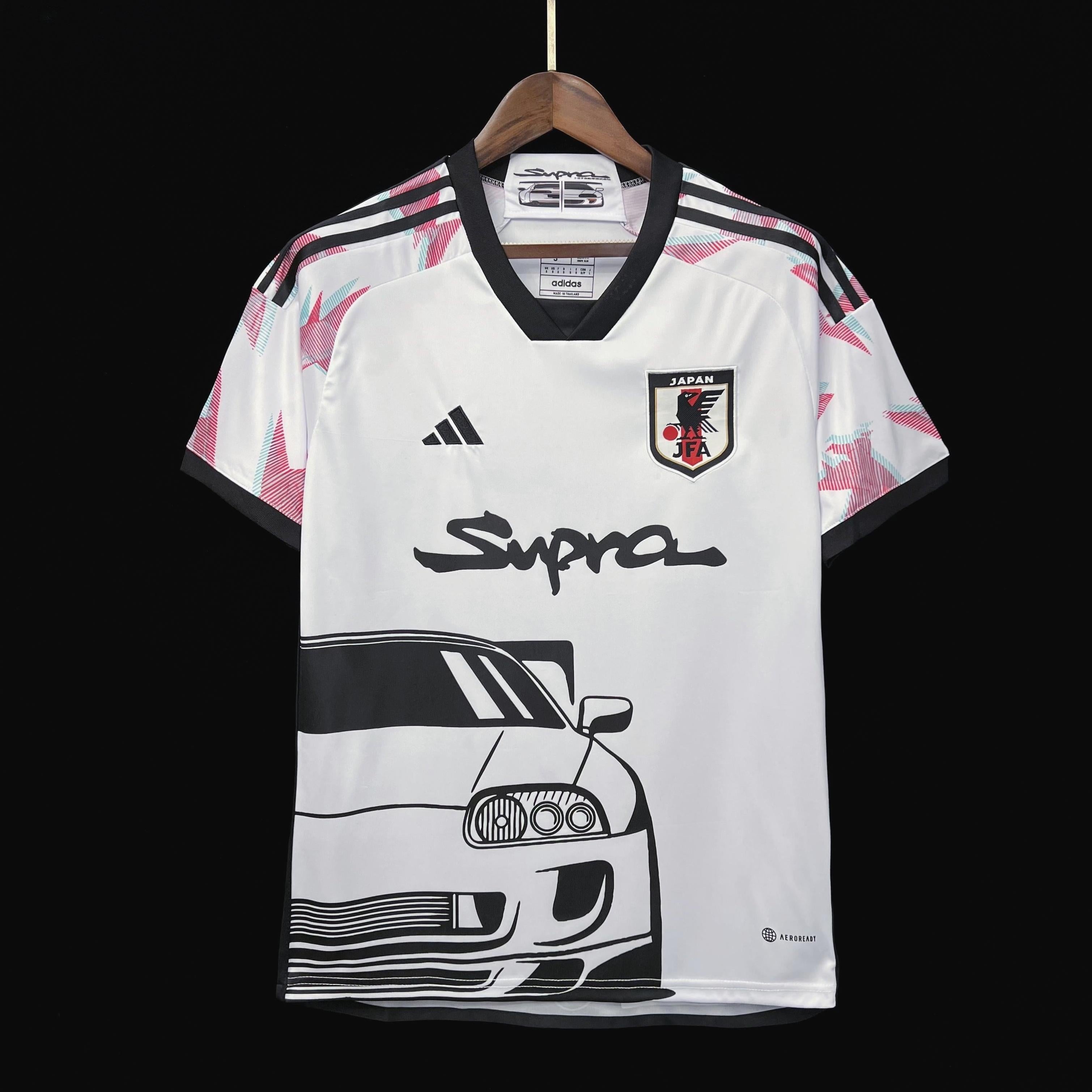 The Retro1999 Japan 2024 Special Edition white football jersey, with black and pink geometric accents, a printed black sports car, and Supra on the front, hangs on a wooden hanger against a black background.