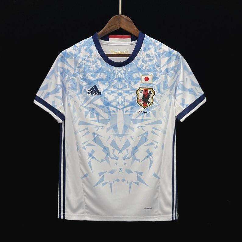A Japan 2024 Special Edition soccer jersey by Retro1999 in white and light blue hangs on a wooden hanger, featuring the Japanese football crest and a geometric blue pattern on the chest and shoulders.
