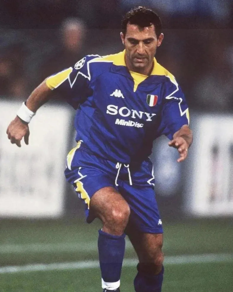 A soccer player in the Retro1999 Juventus 1995-96 Away Shirt, featuring the classic blue and yellow design with SONY MiniDisc sponsor, runs on the field, capturing the spirit of a legendary Champions League Final kit.