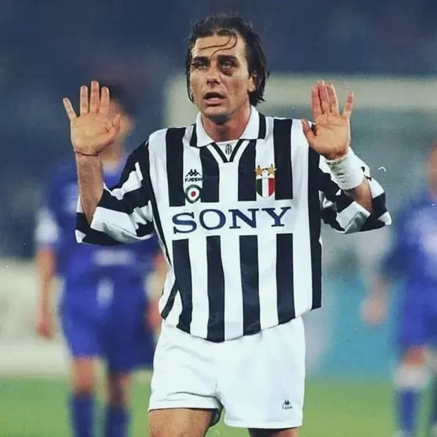 A soccer player wears the Retro1999 Juventus 1995-97 Home Shirt, featuring black and white stripes and “SONY” on the front, standing on the field with hands raised against a backdrop of players in blue kits.