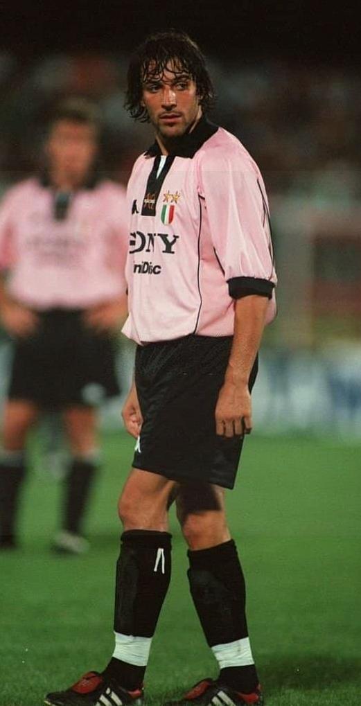 A male soccer player wearing the Retro1999 Juventus 1997-98 Away Shirt stands on the field looking to the side, with another player in a similar kit blurred in the background.