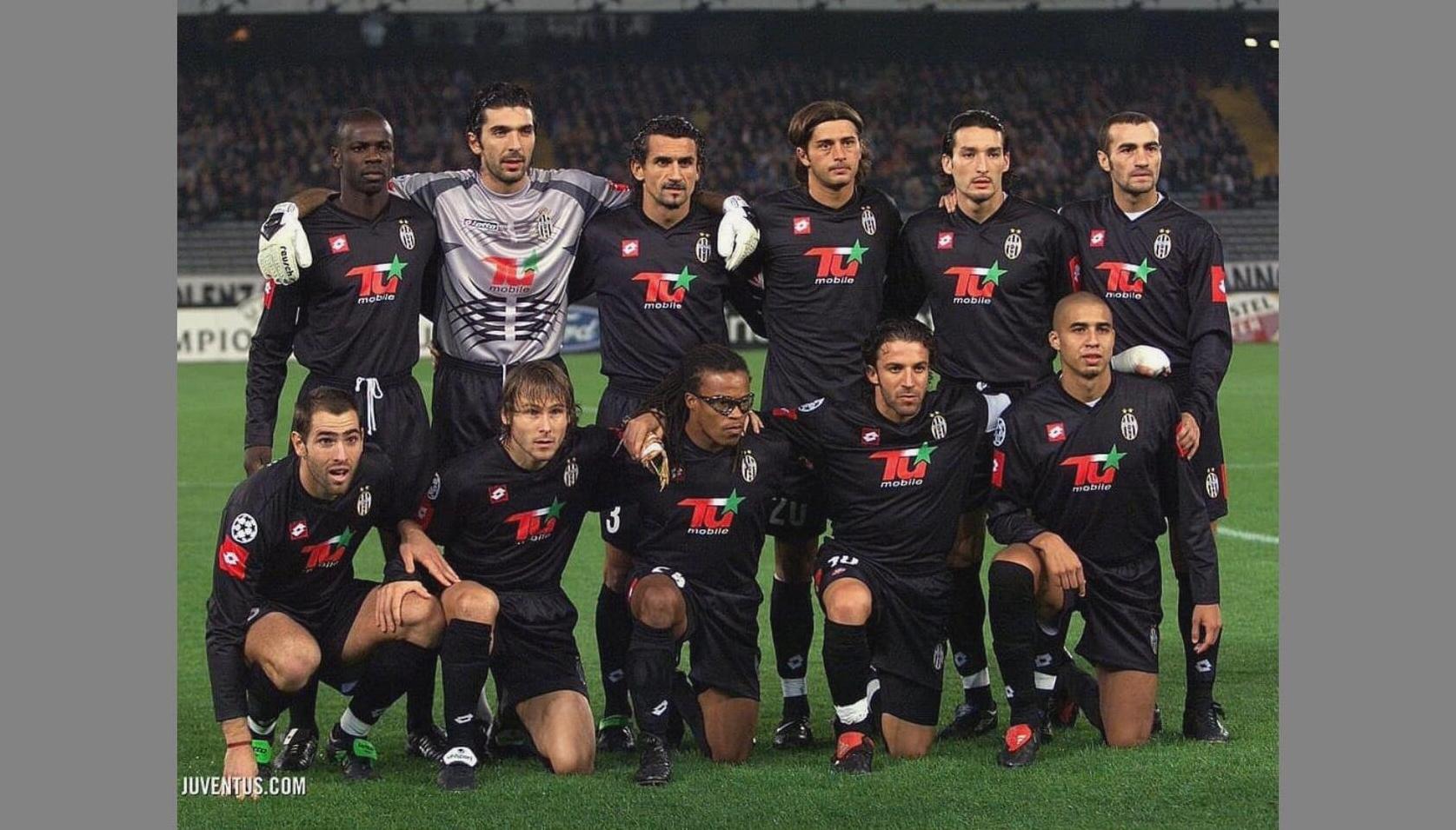 A soccer team in black Retro1999 Juventus 2001-02 Away Shirts poses for a group photo on the field, with seven players standing and four kneeling before a match in a stadium at night.