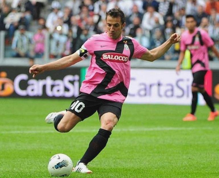 A soccer player wears the Retro1999 Juventus 2011-12 Away Shirt, number 10, and kicks a ball on a green field during a match, with spectators and advertising boards in the background.