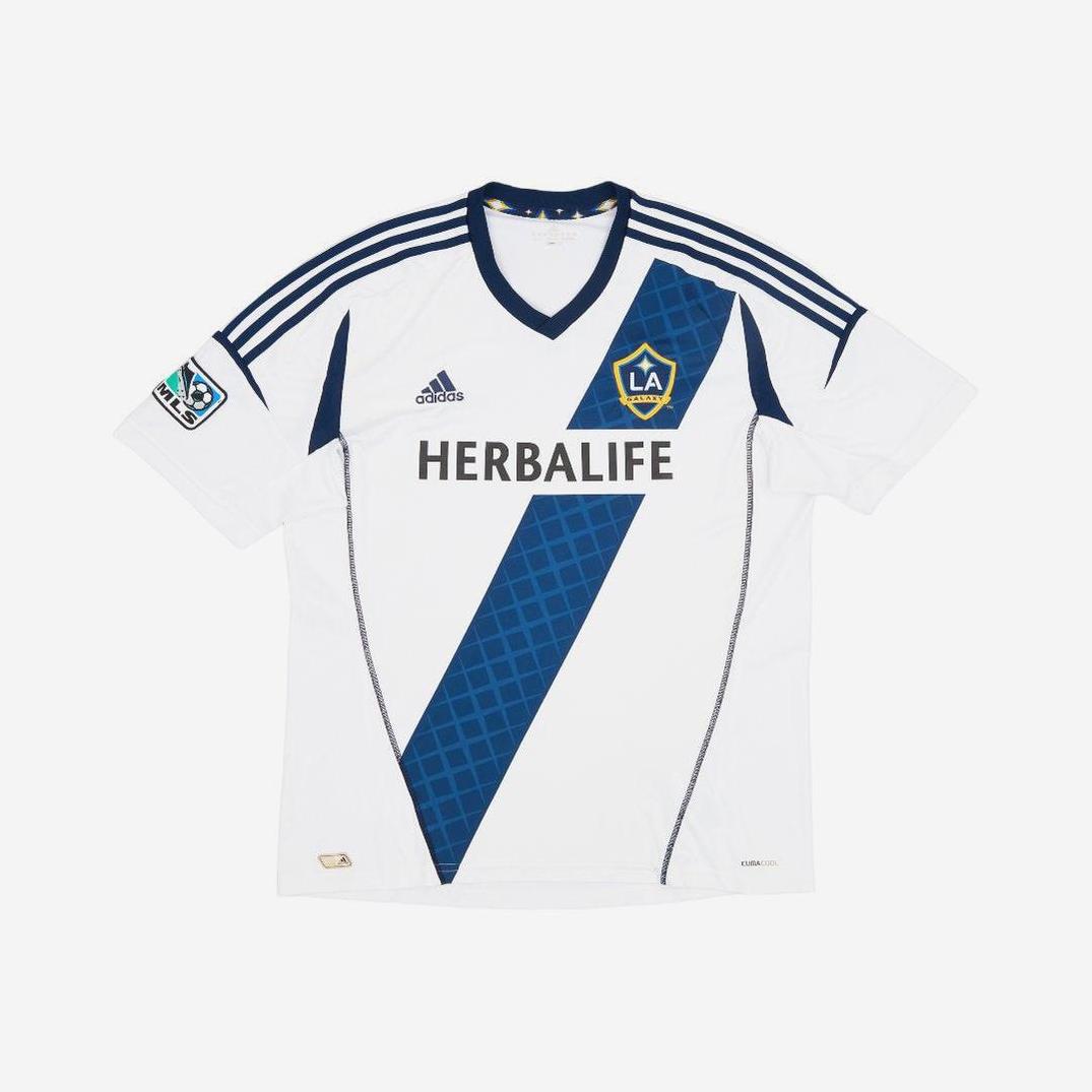 The LA Galaxy 2012-13 Home Shirt by Retro1999 features a white design with a blue diagonal stripe, Adidas logo, Herbalife sponsor, LA Galaxy badge on the chest, and MLS patch—iconic from David Beckham’s last MLS Cup season.