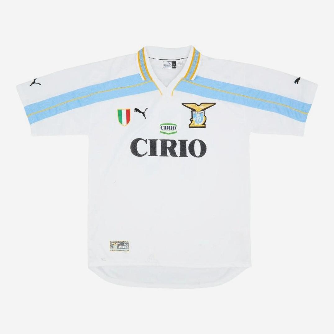 The Retro1999 Lazio 1999-00 Home Shirt is white with light blue and yellow accents, featuring the Puma logo, club crest, CIRIO sponsor, and Scudetto patch. This classic Serie A kit is shown laid flat on a white background.