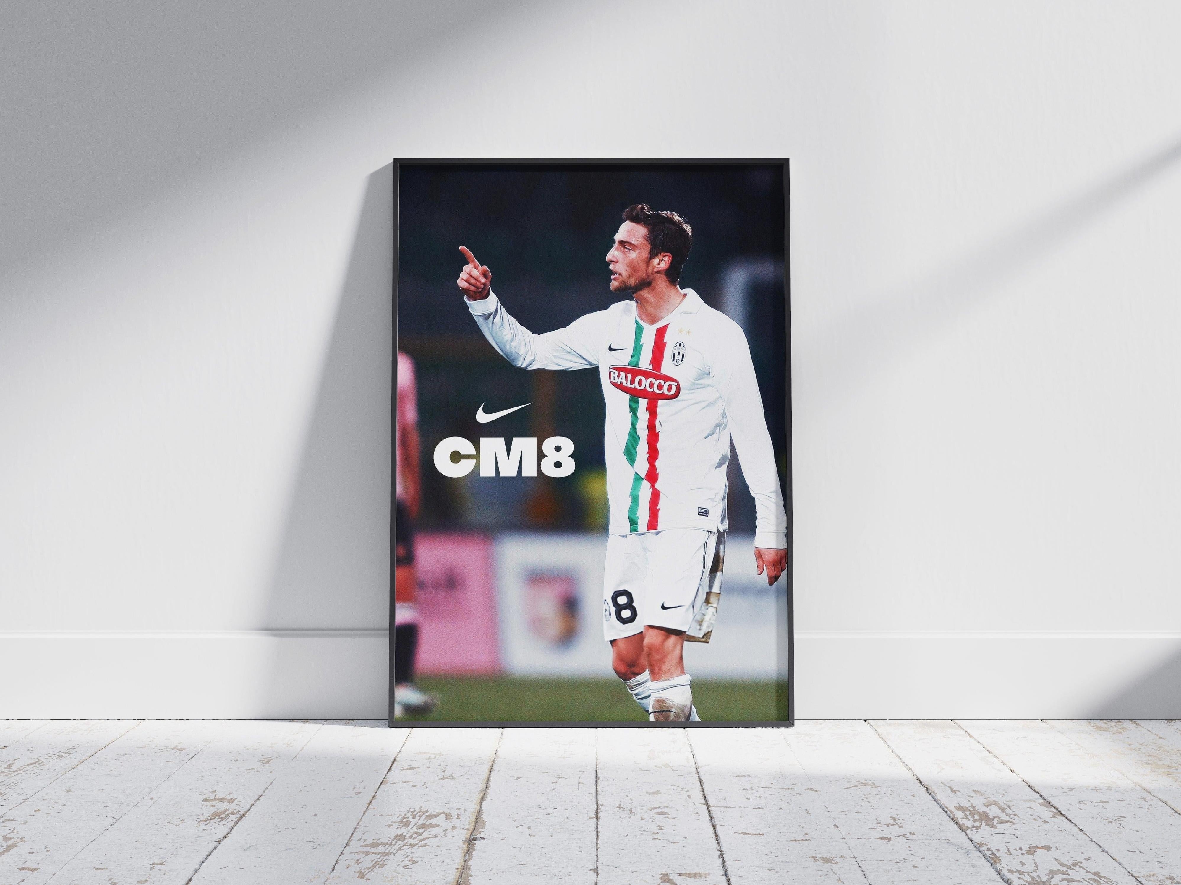 The Marchisio CM8 Poster by Retro1999 features the soccer star in a white Juventus jersey (#8), pointing with his right hand, plus CM8 and Nike logos. Framed, it rests against a white wall—ideal decor for football fans.