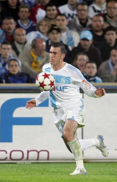 Wearing the Retro1999 Marseille 2005-06 Home Shirt, a soccer player sprints across the field, focused on a red and white ball, as fans watch from the stands.