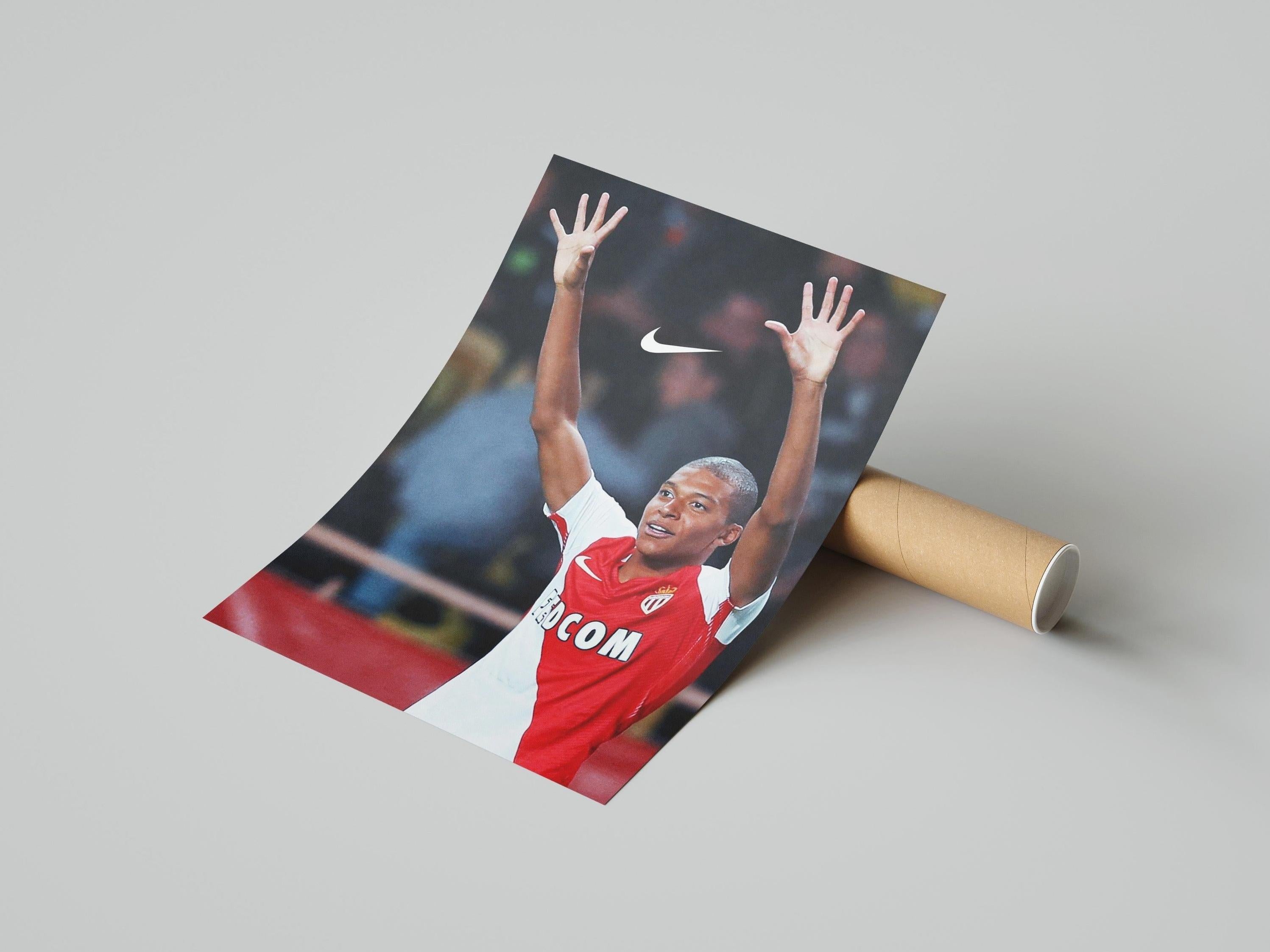 The Mbappe x AS Monaco Poster by Retro1999 shows a soccer player in a red and white jersey with arms raised in celebration, featuring the Nike logo above him. The poster is rolled up and displayed on a flat surface.