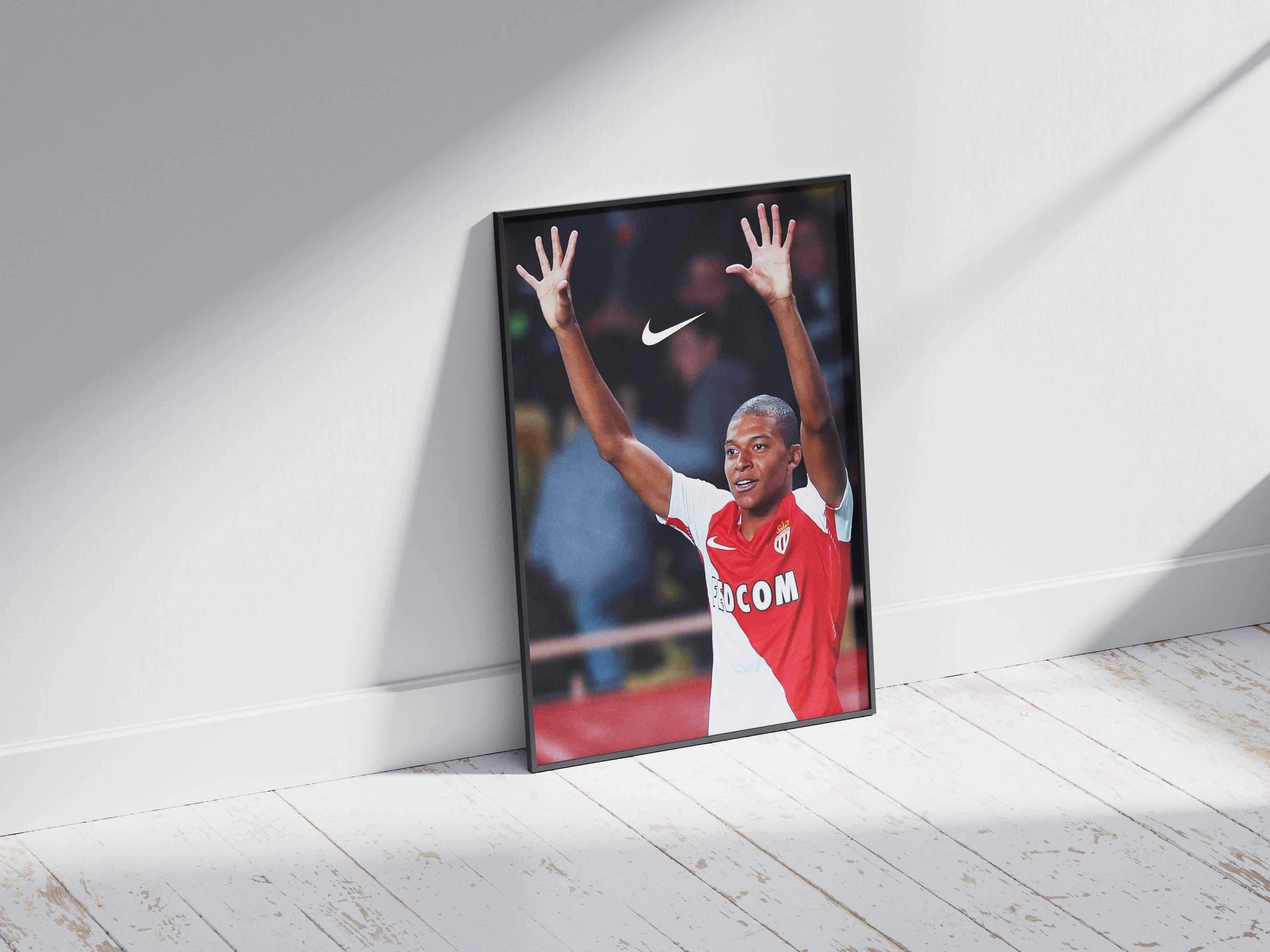 A framed Mbappe x AS Monaco Poster by Retro1999 leans against a white wall, showing the soccer star in a red and white jersey with raised arms celebrating. The Nike logo appears above him as sunlight casts shadows on the floor.