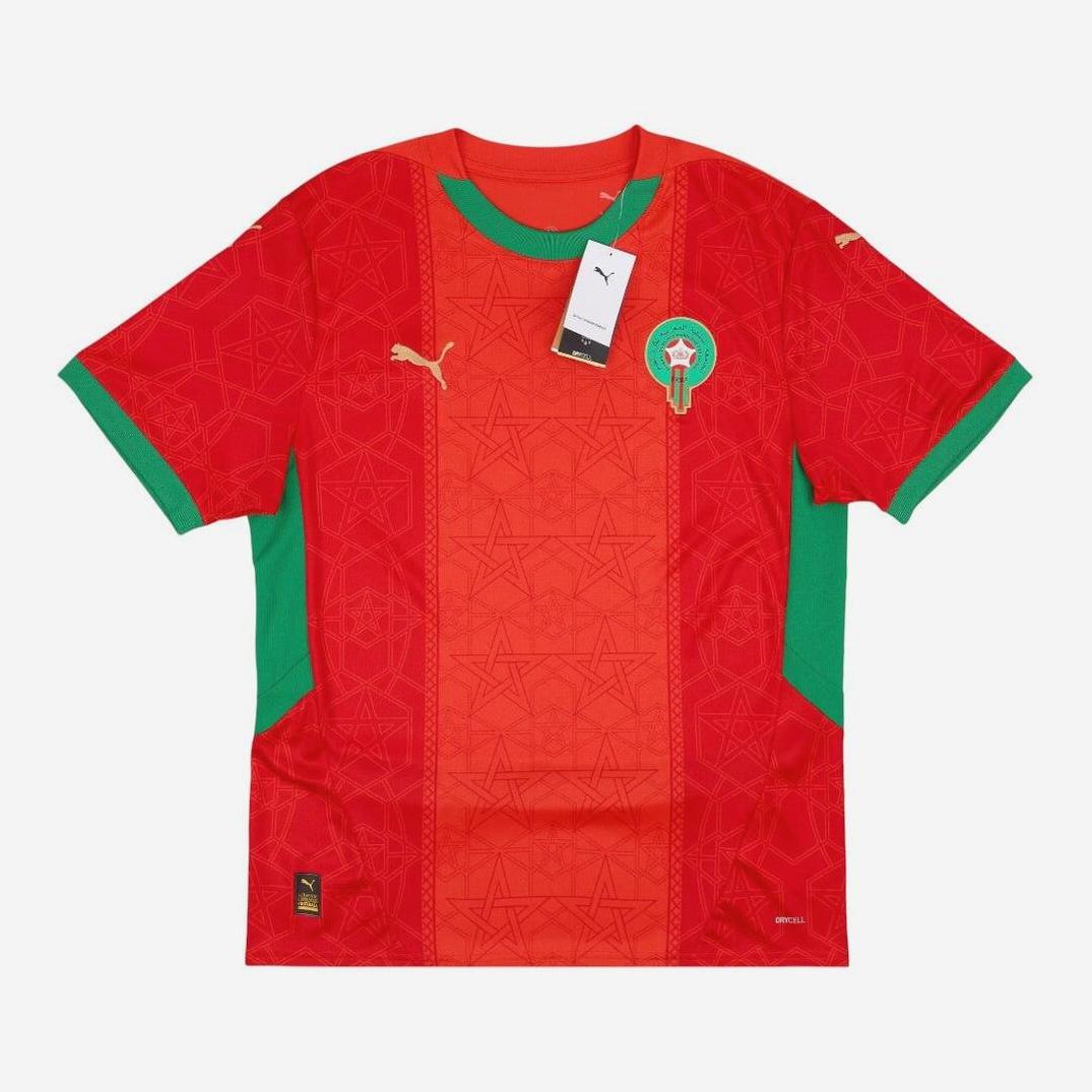 The Morocco 2024-25 Home Shirt by Retro1999 is red with green side panels, gold Puma logos, and the team crest on the chest. Tags are attached to the collar, and the retro design features subtle star patterns.