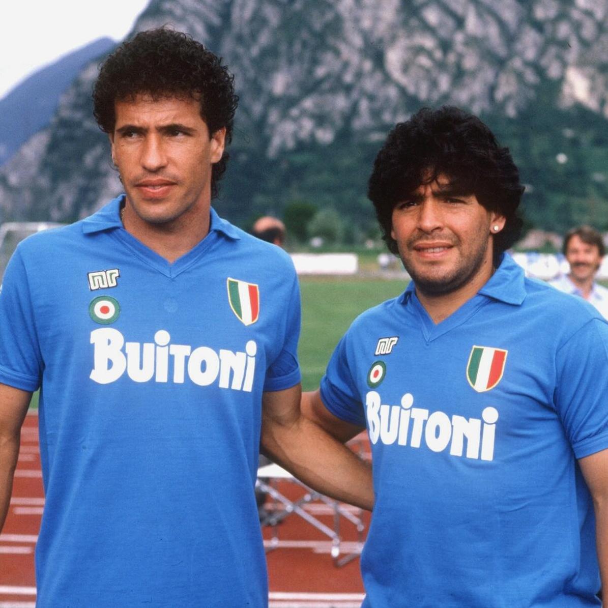 Two men in Retro1999 Napoli 1987-88 Home Shirts stand outdoors with mountains and greenery behind them. Both have dark, curly hair and look relaxed and friendly in this classic retro football kit.