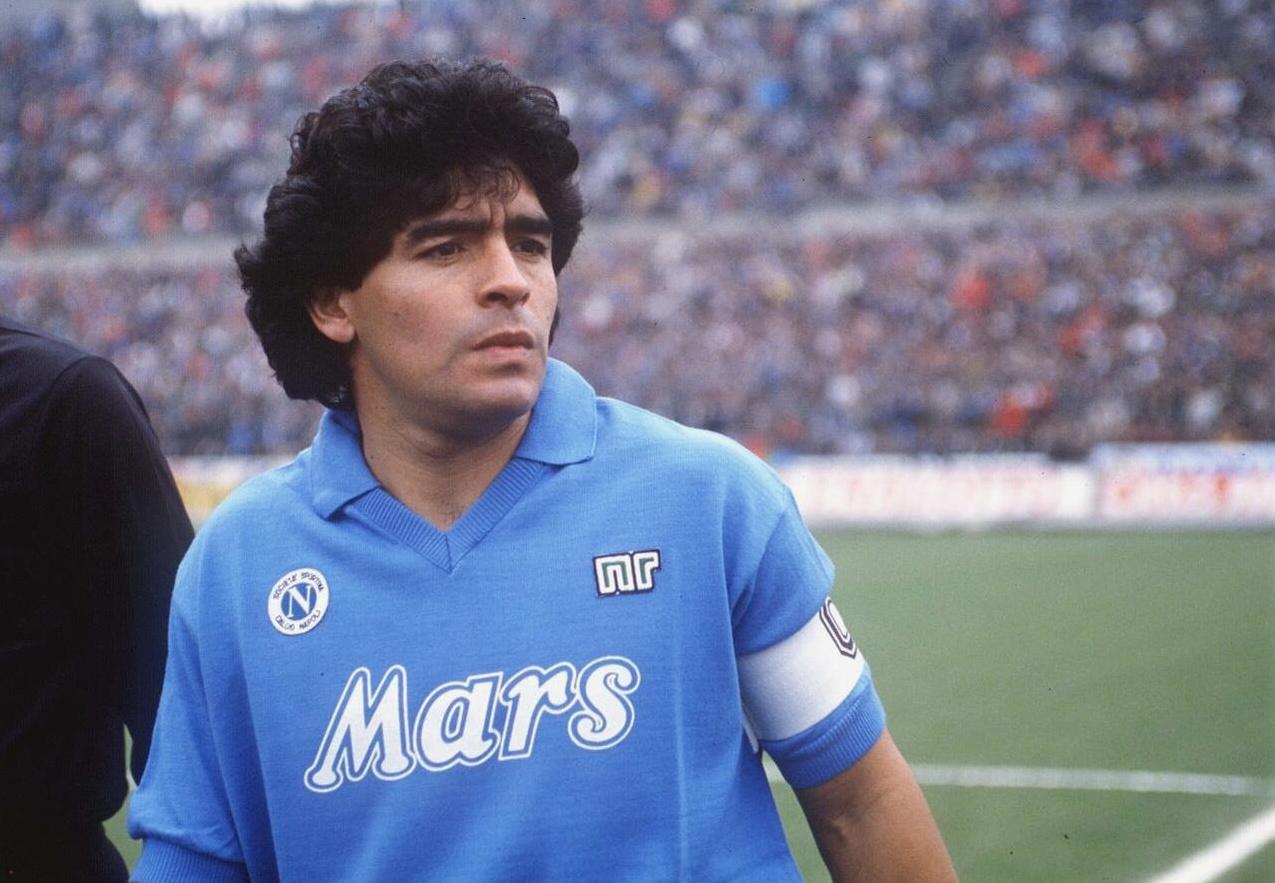 A male soccer player wears the Retro1999 Napoli 1988-89 Home Shirt with Mars sponsor and captain’s armband, standing on the field like Maradona during their UEFA Cup win, a blurred stadium crowd behind him.