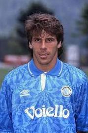 A young man with brown hair wears the Retro1999 Napoli 1991-93 Home Shirt, featuring a blue pattern, white collar, and Voiello sponsor. He stands outdoors on a field with blurred trees in the background—ideal for football fans.