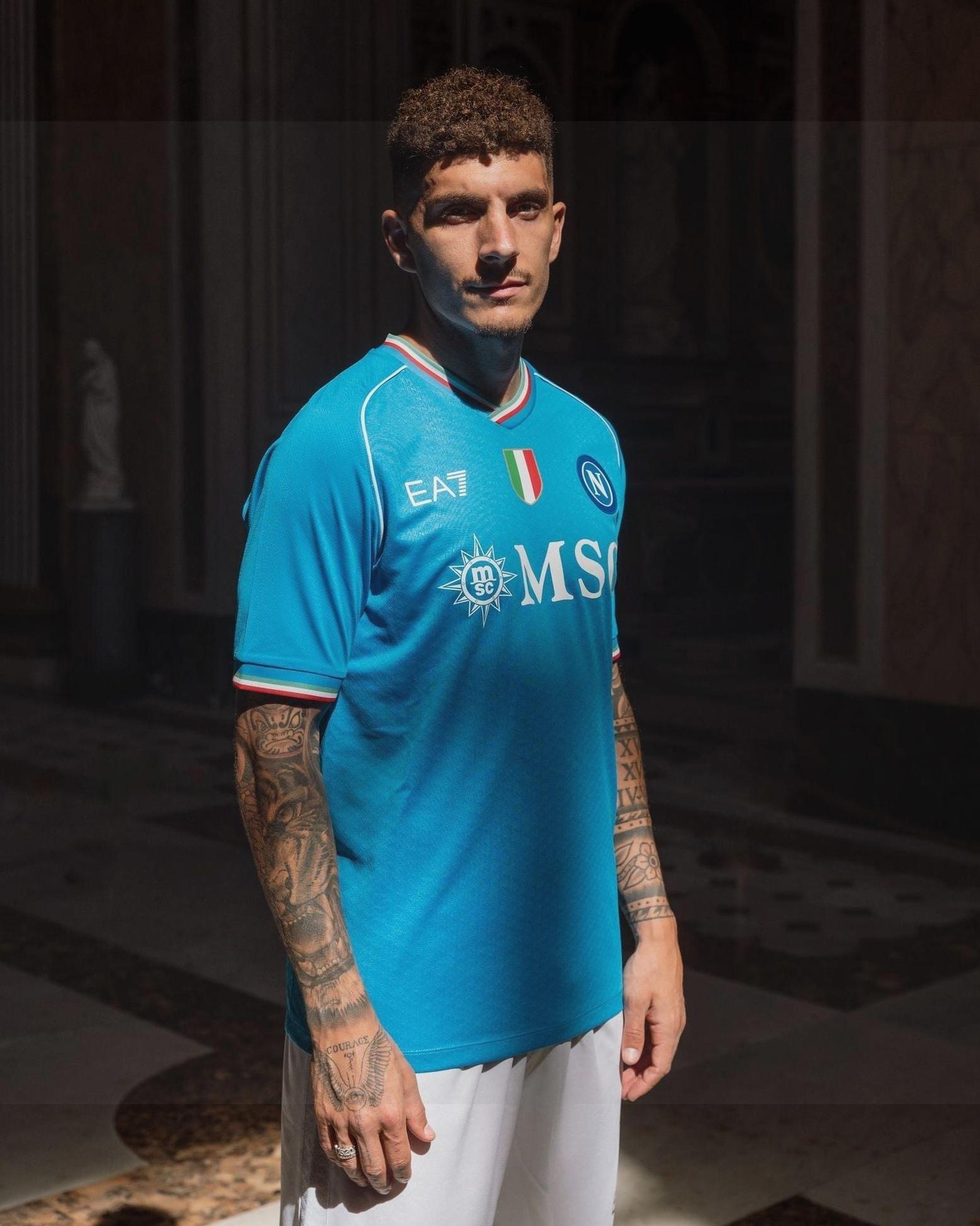 A man with short curly hair and tattoos models the Retro1999 Napoli 2023-24 Home Shirt and white shorts in a dimly lit room, representing true Napoli passion.