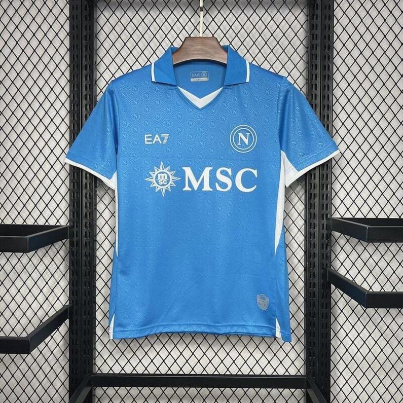 The Retro1999 Napoli 2024-25 Home Shirt in blue with white trim hangs on a wooden hanger against a black mesh background, displaying the club’s logo, EA7, and MSC sponsor logos on the front.