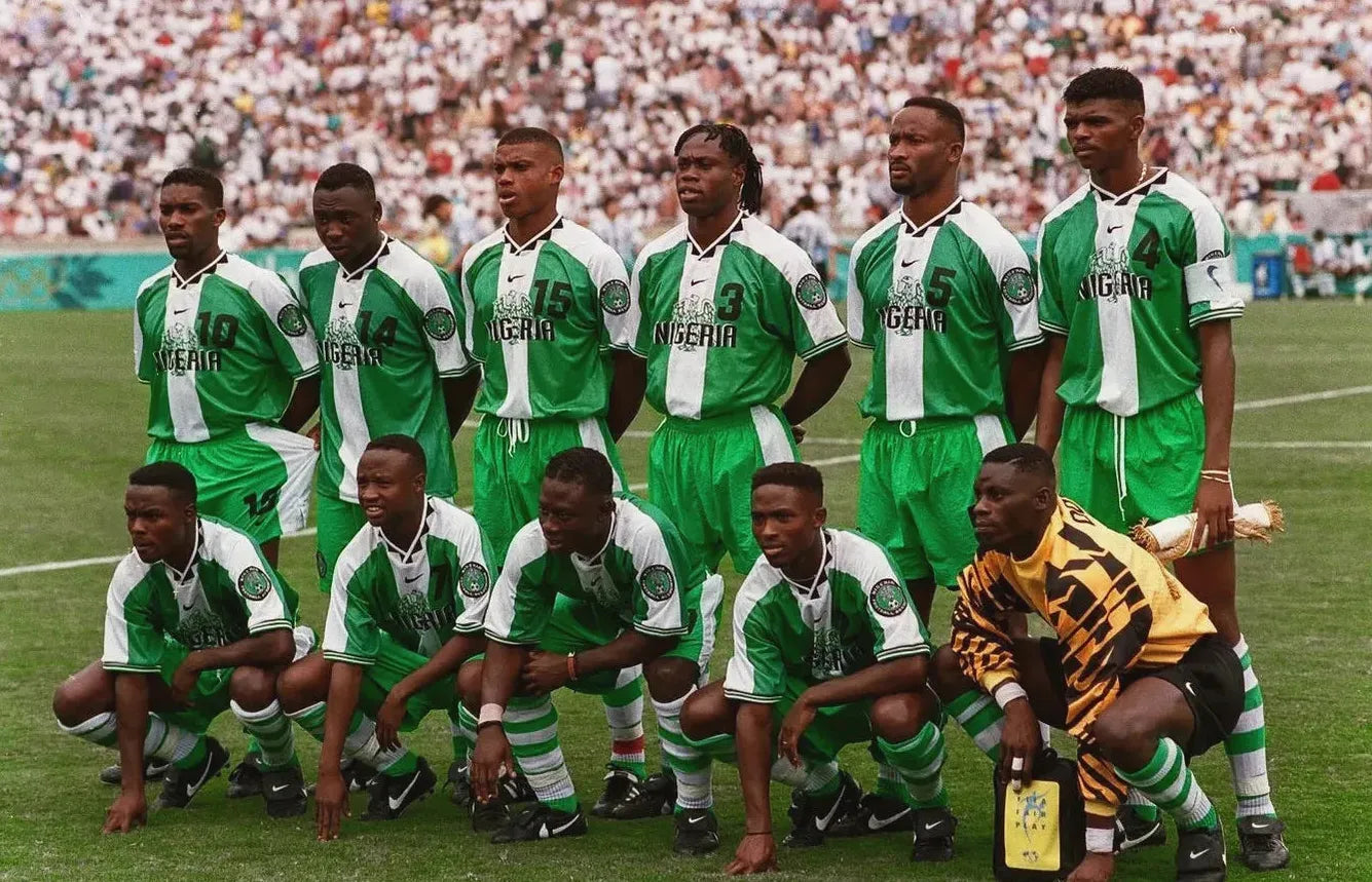 Eleven Super Eagles players wear the Retro1999 Nigeria 1996 Home Shirt for a team photo before a match, with a packed stadium behind them. The goalkeeper is distinct in yellow and black at the African Cup of Nations.