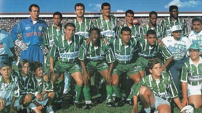 A soccer team in green and white stripes, wearing the Retro1999 Palmeiras 1996 Home Shirt, poses on the field with coaches and young mascots as fans fill the stadium in the background.