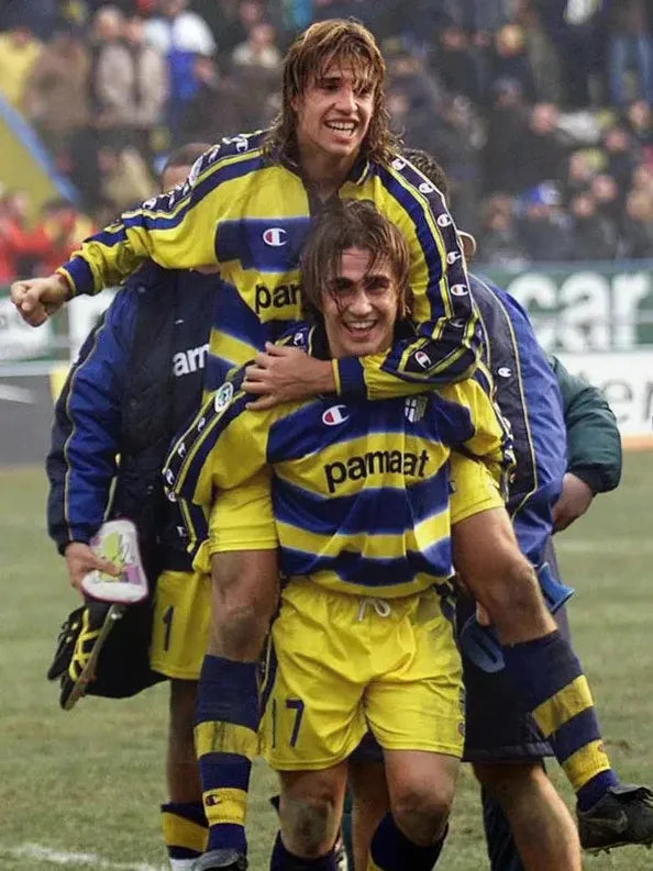 Two soccer players in yellow and blue Retro1999 Parma 1999-00 Home Shirts celebrate on the field, one carrying the other on his back with smiles, as blurred teammates and spectators capture this classic Serie A club moment.