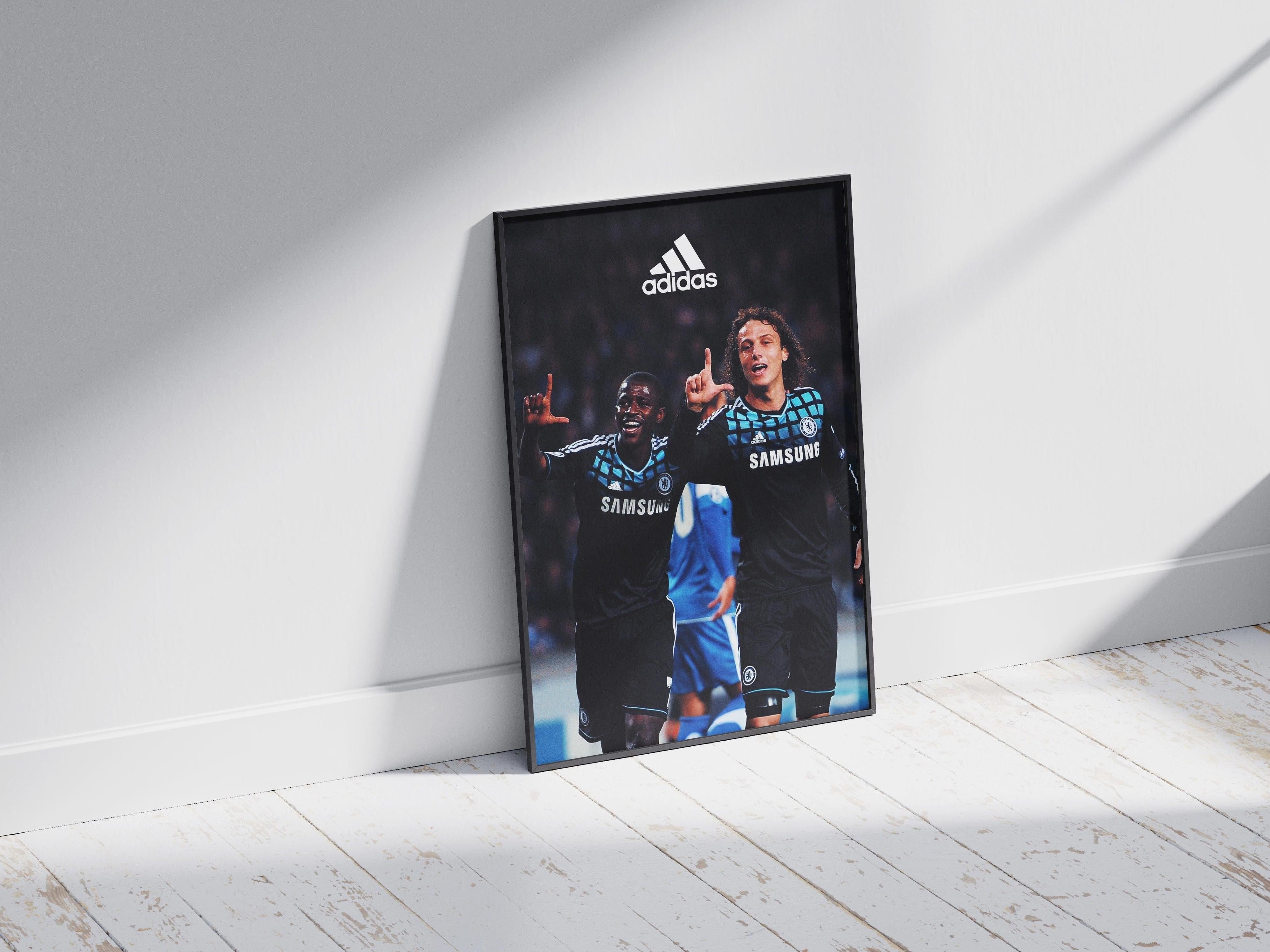 The Ramires x David Luiz Poster by Retro1999 shows the two Chelsea FC players in black kits celebrating on the field, with the Adidas logo above them. The framed print stands against a white wall on a sunlit wooden floor, perfect for football fans.