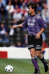 A soccer player wears the Retro1999 Rangers F.C. 1994-95 Third Shirt, dribbling a white ball on the field with blurred spectators behind—a stylish tribute to football history.