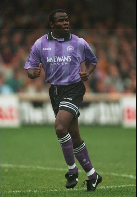 A footballer wears the Retro1999 Rangers F.C. 1994-95 Third Shirt with McEwans Lager sponsor, black shorts, and socks, sprinting on a grassy field during a match as fans fill the stands—a true retro football scene.