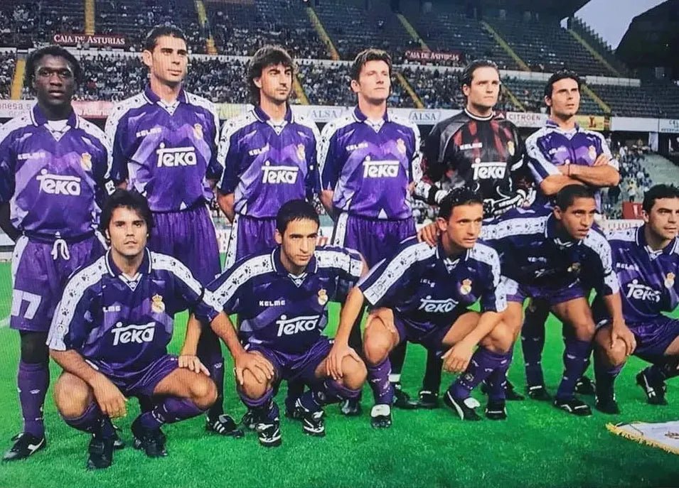 A soccer team in the Retro1999 Real Madrid 1996-97 Away Shirt poses for a group photo on the stadium field, with stands and spectators in the background—celebrating classic football history.