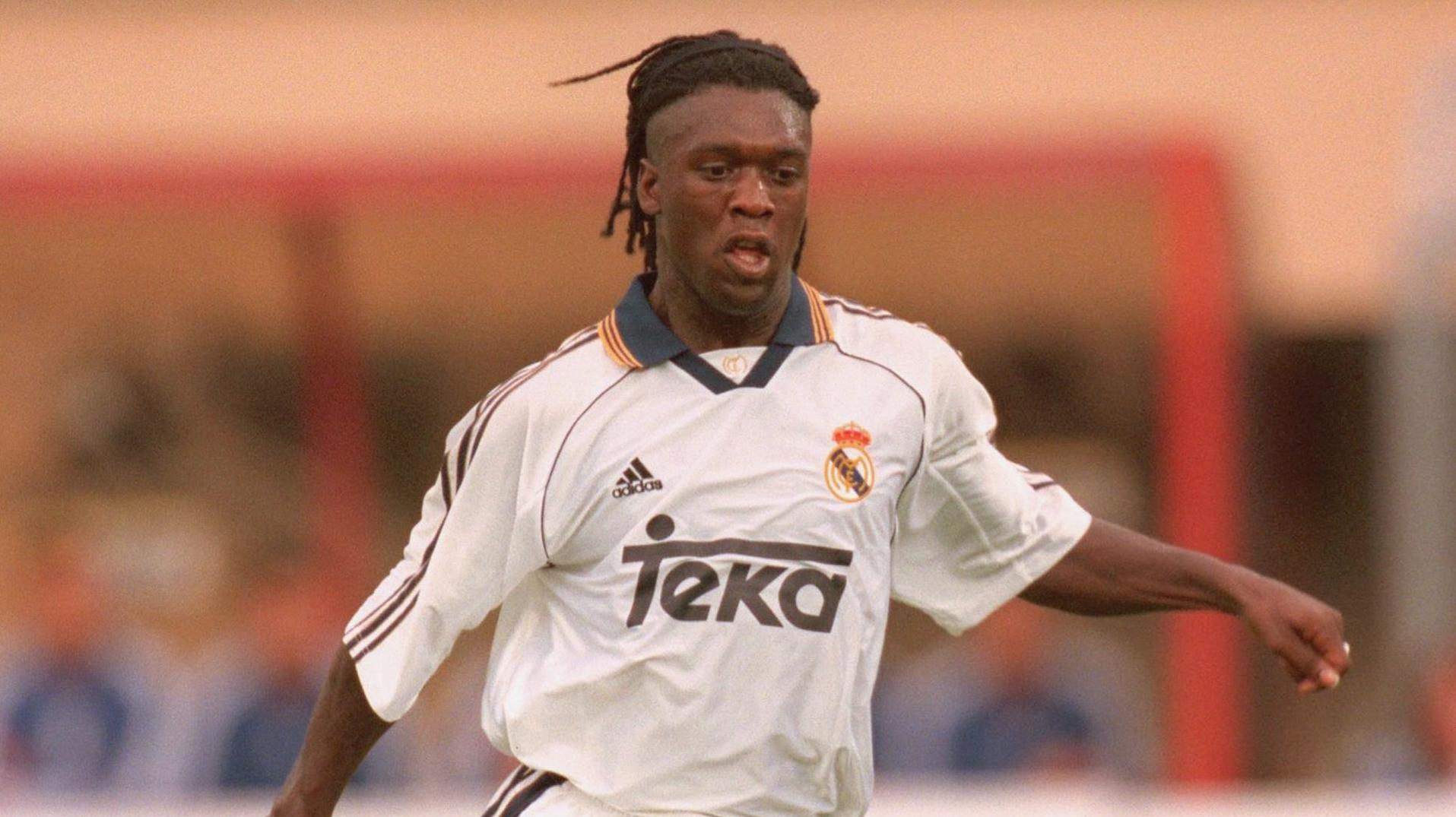 A soccer player in a Retro1999 Real Madrid 1999-00 Home Shirt, featuring the Teka sponsor and Adidas logo, runs on the field with short dreadlocks, highlighting the shirt’s classic retro design during gameplay.