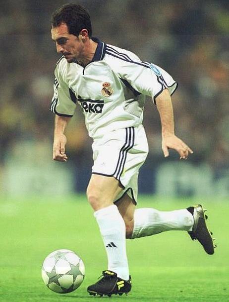 A soccer player in a Retro1999 Real Madrid 2000-01 Home Shirt and black cleats dribbles a ball across the green field, focused and skillful, with fans blurred in the background, evoking classic La Liga style.