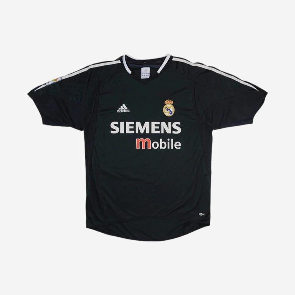 The Real Madrid 2004-05 Away Shirt by Retro1999 features a black design with white trim, Siemens Mobile sponsor, Adidas logo, and club crest—a must-have retro kit for fans and collectors.