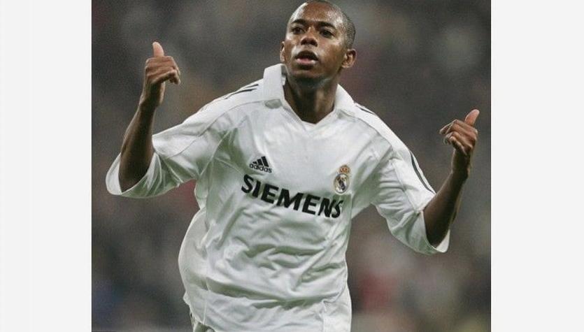 A soccer player wears the Real Madrid 2005-06 Home Shirt by Retro1999, raising both thumbs and looking upward during a match. The blurred background highlights the iconic Siemens-fronted retro kit, drawing focus to the player.