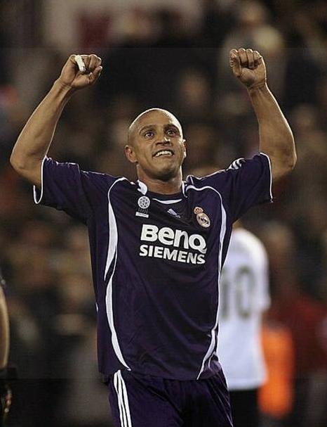 A soccer player wearing the Retro1999 Real Madrid 2006-07 Third Shirt with BenQ Siemens sponsor celebrates on the field, arms raised, with a blurry crowd and an opposing player in the background.