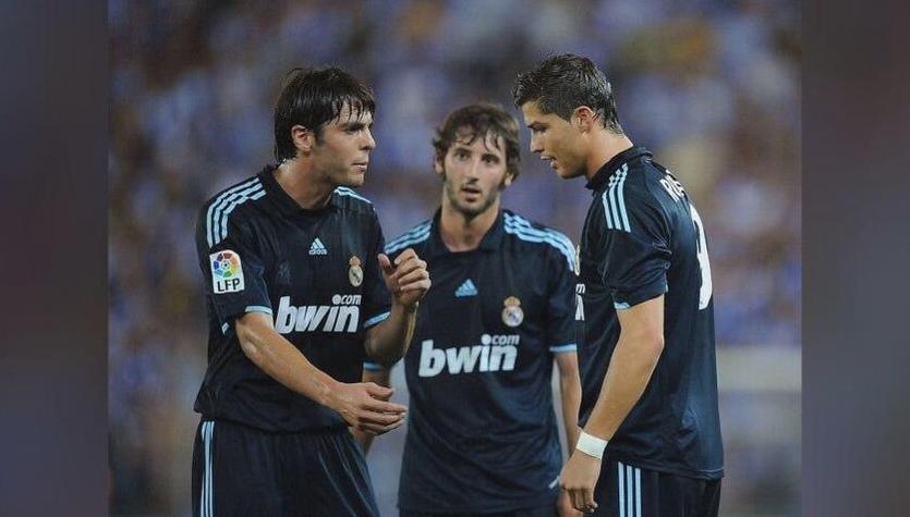 Three soccer players wear the Retro1999 Real Madrid 2009-10 Away Shirt as they stand on the field, seemingly discussing tactics. The stadium background is blurred and filled with fans.