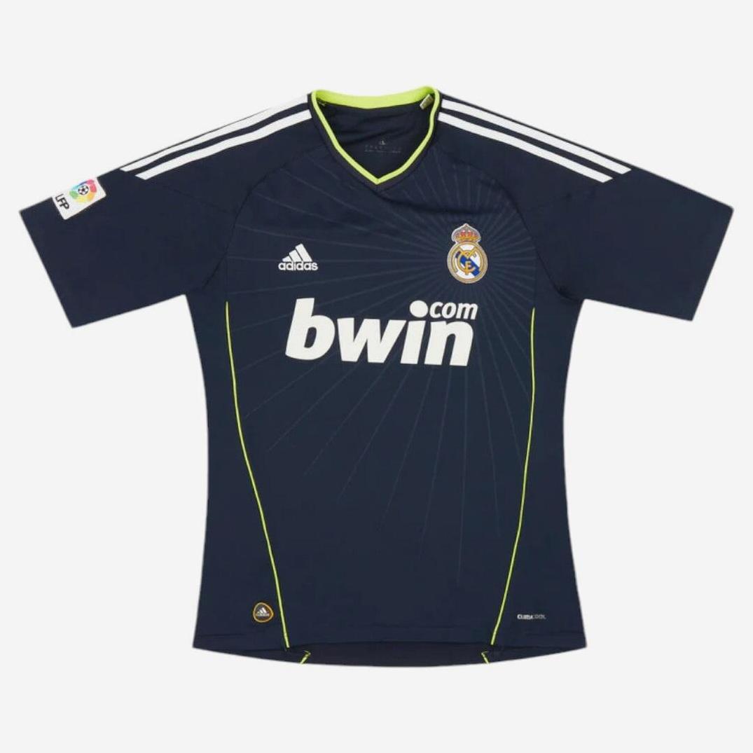 The Real Madrid 2010-11 Away Shirt by Retro1999 is navy blue with white shoulder stripes, yellow piping, Adidas logo, club crest, bwin.com sponsor, and a La Liga patch, displayed flat on a white background.