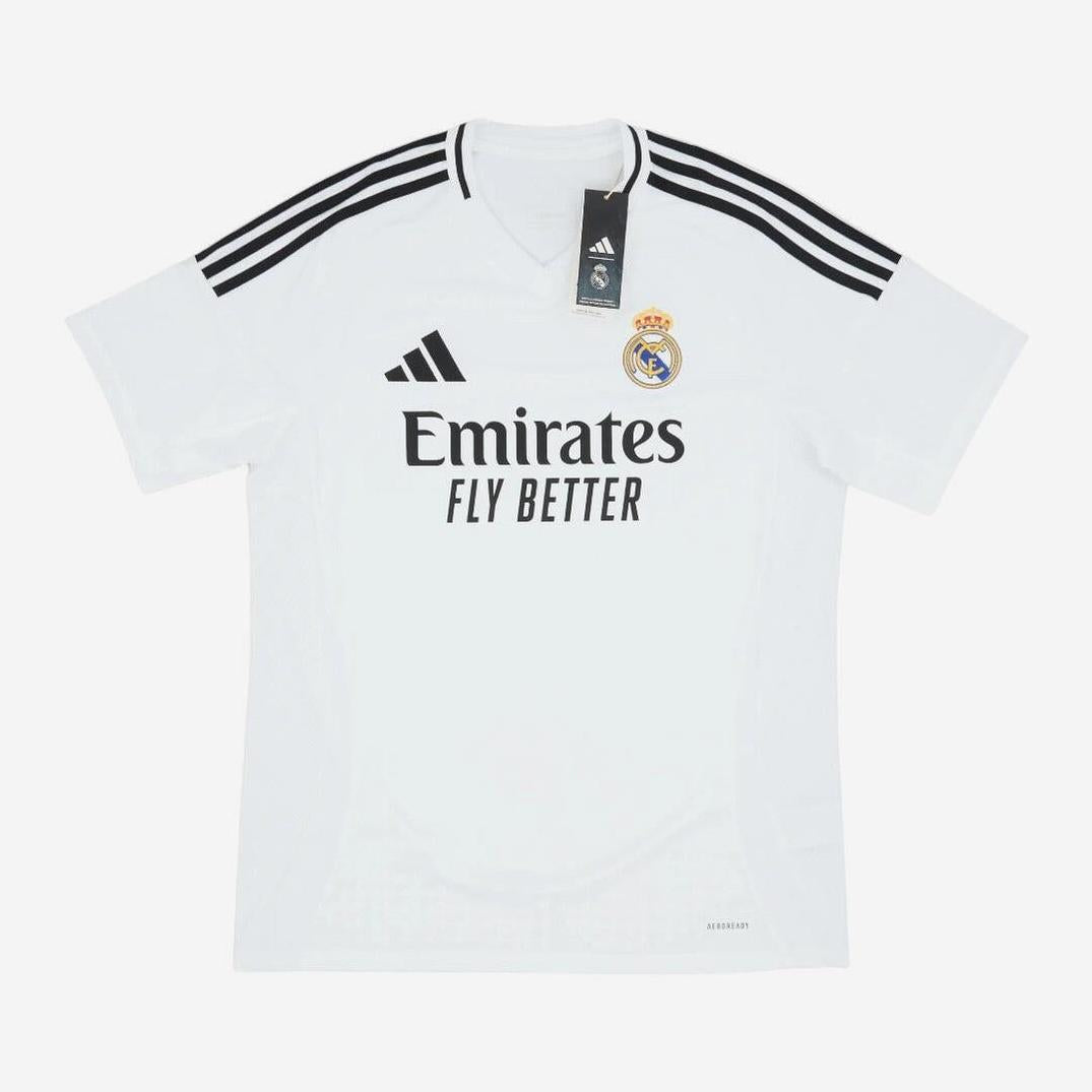 The Retro1999 Real Madrid 2024-25 Home Shirt is white with black shoulder stripes, club crest, Emirates Fly Better sponsor, and classic styling—ideal for fans. A black tag is attached to the collar.