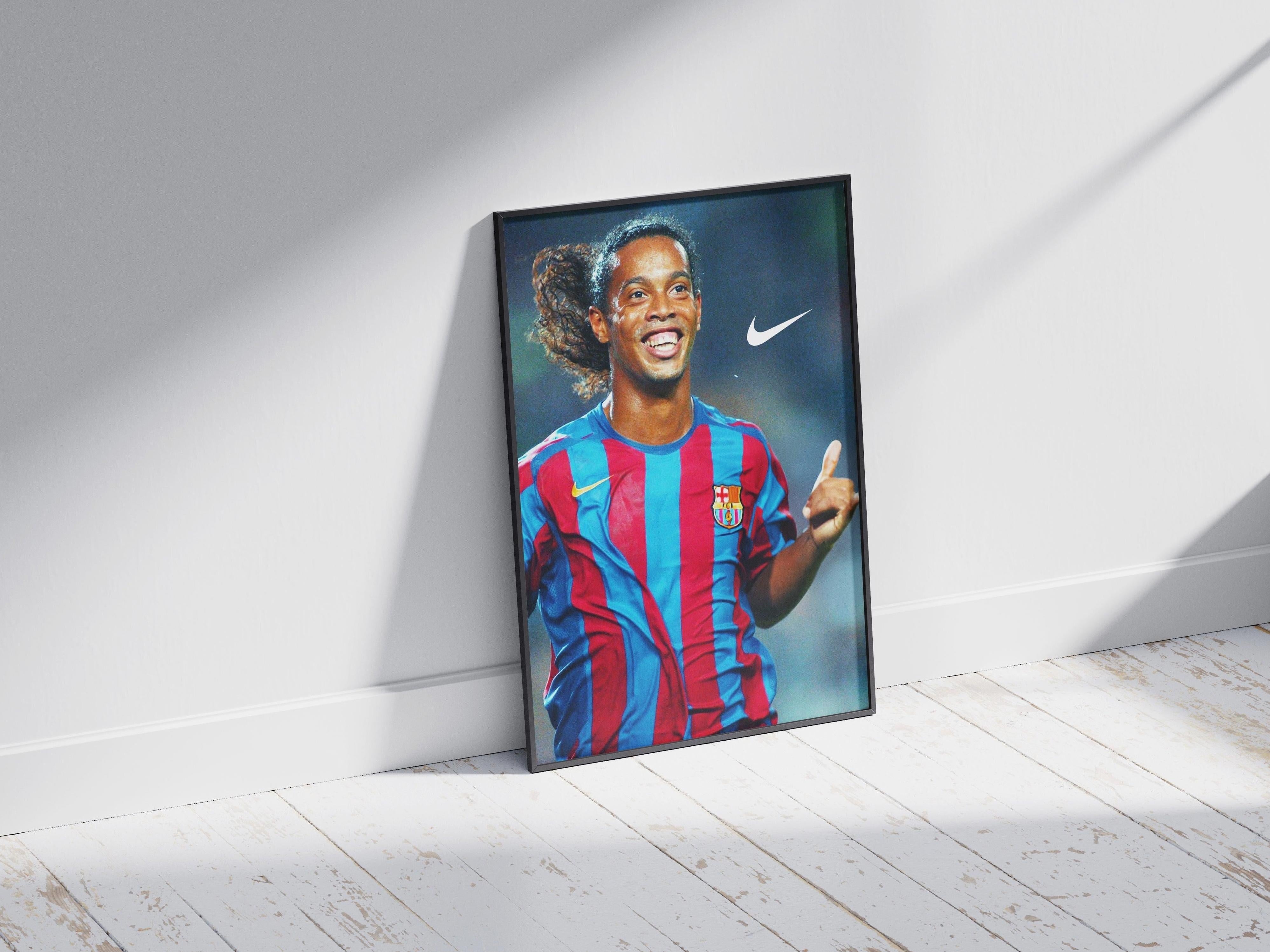 The Ronaldinho Gaucho x Barcelona Poster by Retro1999 features the soccer star smiling in a red and blue Barcelona jersey, with sunlight casting shadows—perfect memorabilia for any football fan. The white Nike logo is visible above.