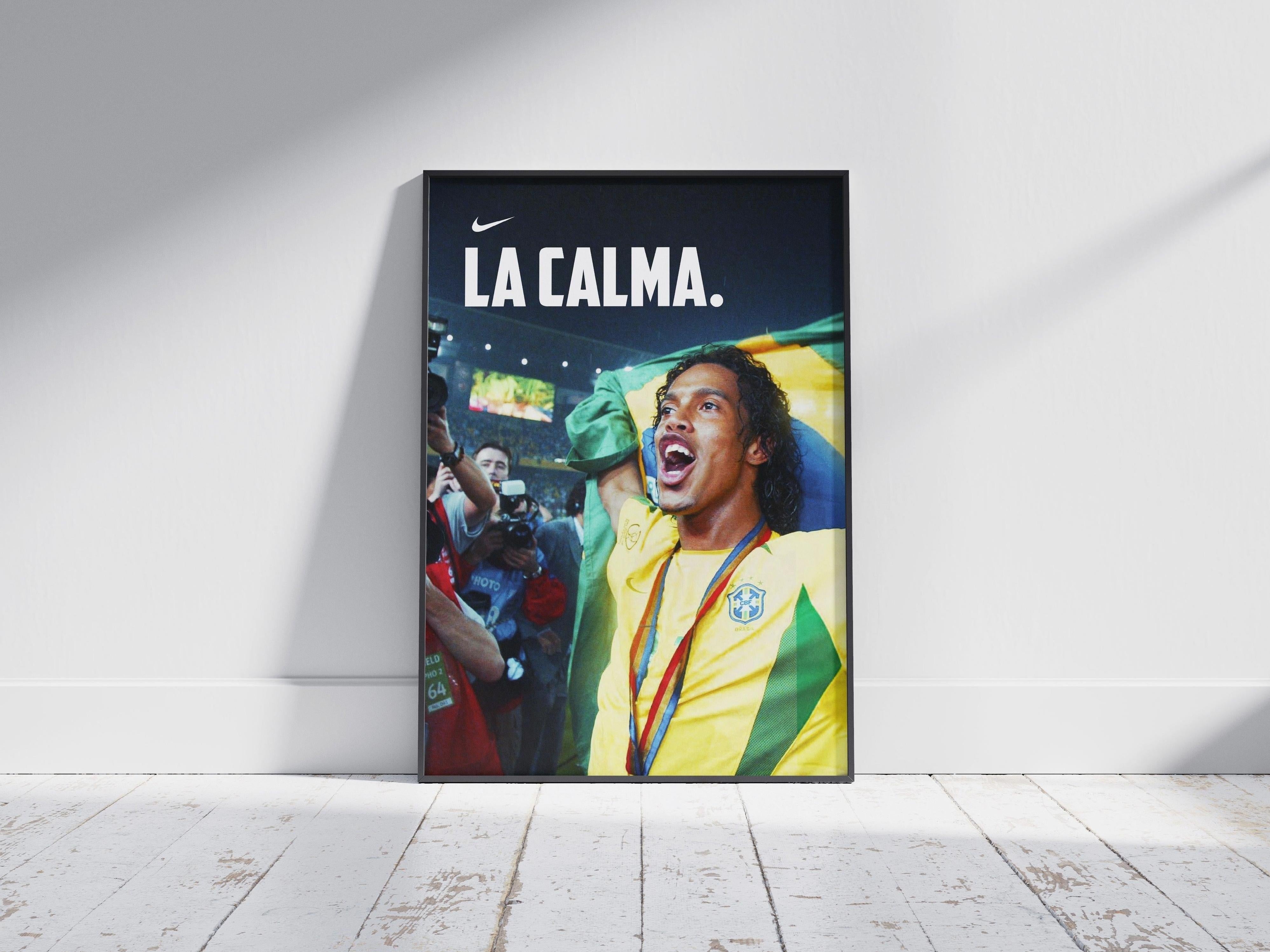 The Retro1999 Ronaldinho La Calma Poster shows the Brazilian soccer star celebrating his 2002 FIFA World Cup win with medal and flag, Nike logo, and LA CALMA. on top, framed and displayed on a wooden floor with light casting a shadow behind.