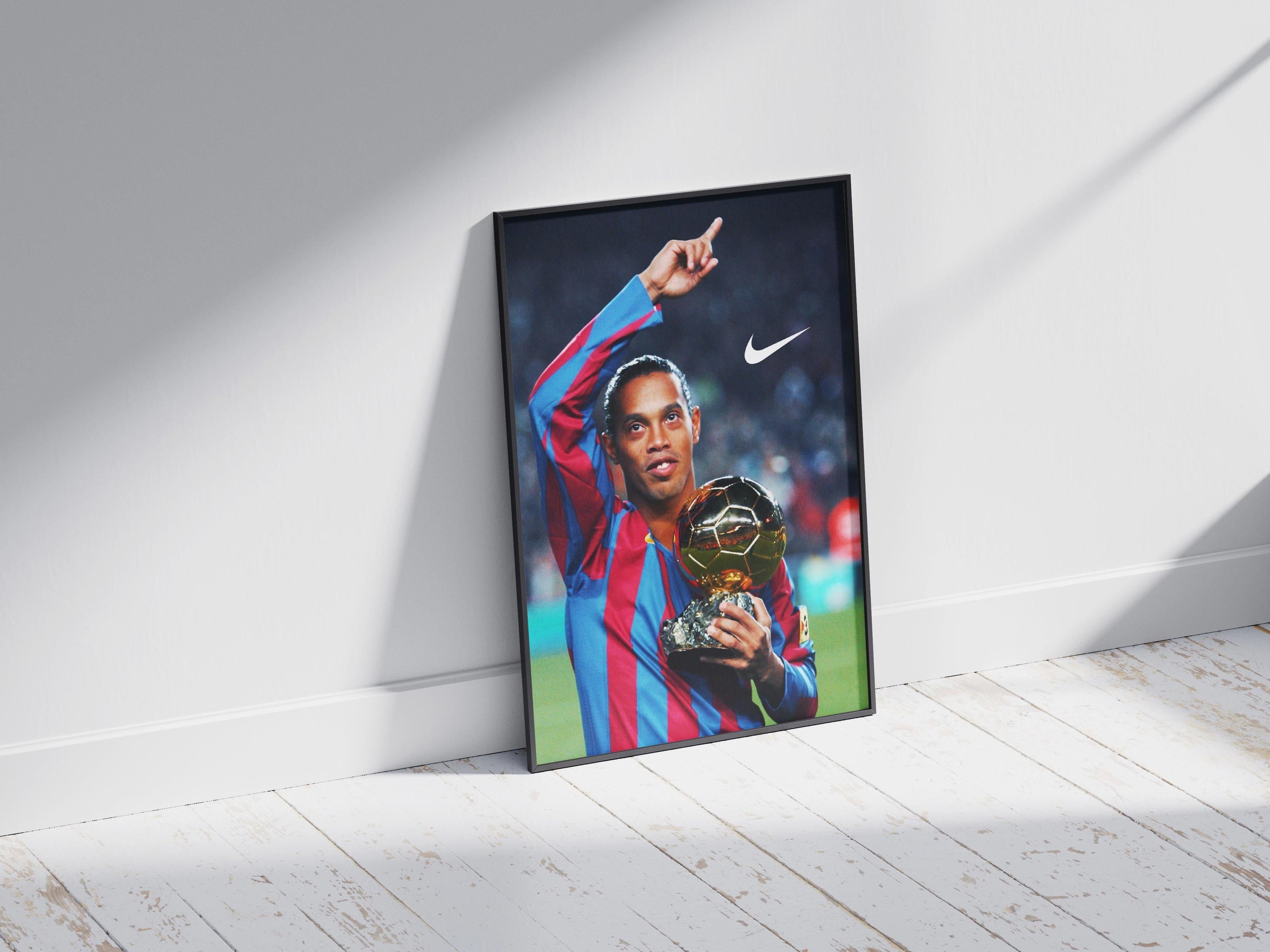 The Ronaldinho x Ballon dOr Poster by Retro1999 features a framed photo of the soccer legend in a red and blue jersey, celebrating with his Ballon dOr trophy. The iconic Nike logo appears in the top right corner, set against a wooden floor backdrop.