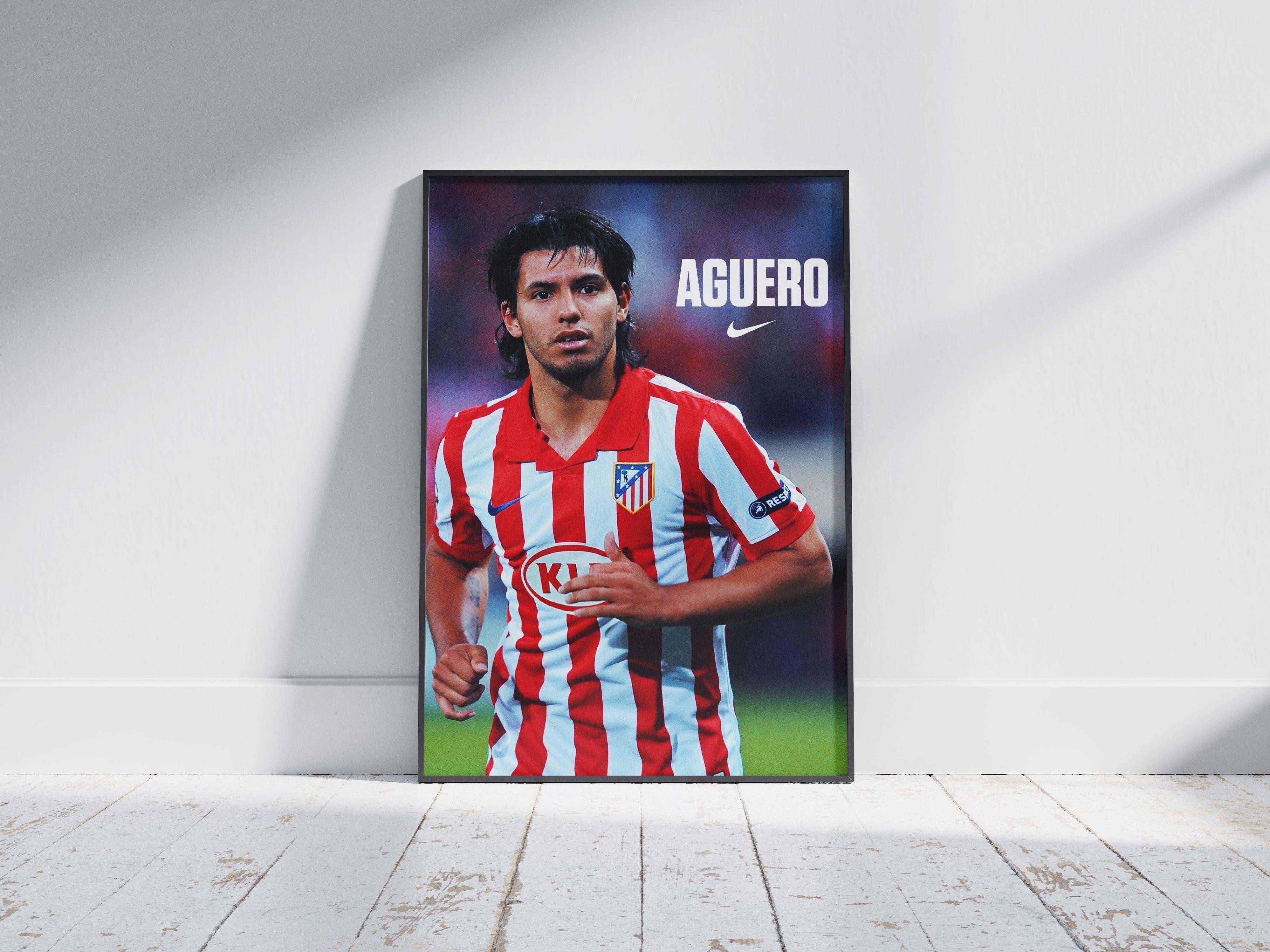 The Sergio Aguero x Atletico Madrid Poster by Retro1999, featuring Aguero in a red and white striped jersey, stands framed on a wooden floor against a white wall—an ideal collectible for football fans or memorabilia collectors.