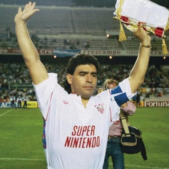 A soccer player in the Retro1999 Sevilla 1991-92 Home Shirt with Super Nintendo sponsor lifts a pennant on a La Liga field, stadium and fans in the background.