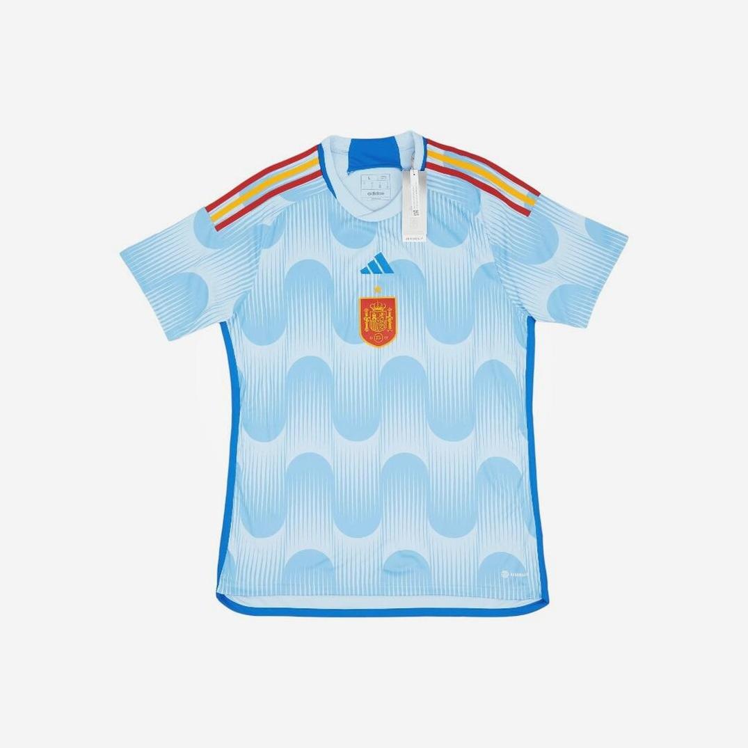 The Spain 2022 Away Shirt by Retro1999 features a light blue design with wavy patterns, red and yellow shoulder stripes, Adidas logo, and Spains crest on the chest. Crafted from breathable materials and shown on a white background.