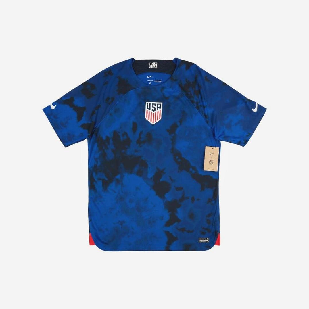 The USA 2022 Away Shirt by Retro1999 is blue with a black tie-dye design, featuring double Nike logos on the sleeves and the team crest on the chest. A beige tag is attached on the right side.