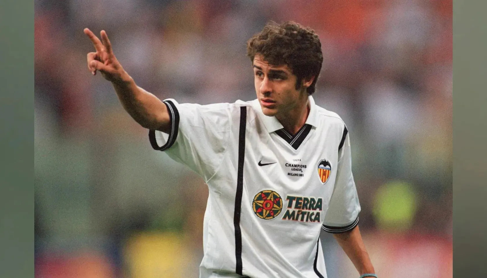 A soccer player in a Retro1999 Valencia CF 2000-01 Home Shirt holds up two fingers with his right hand, standing before a blurred crowd of football fans and the field.