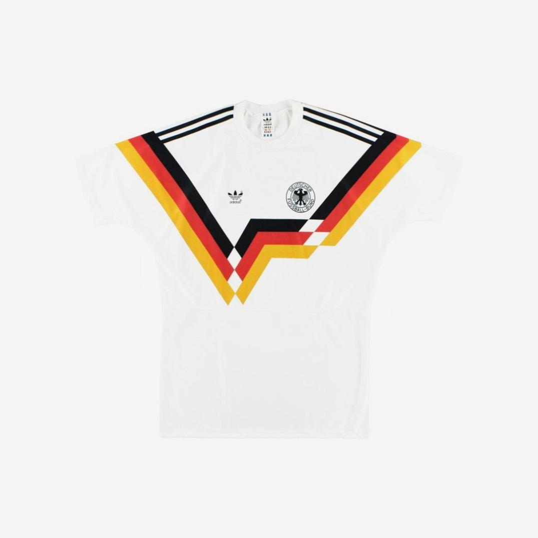 The Retro1999 West Germany 1990 Home Shirt is a white retro kit with black, red, and yellow V-shaped geometric stripes across the chest, featuring a circular crest on the right and the brand logo on the left.